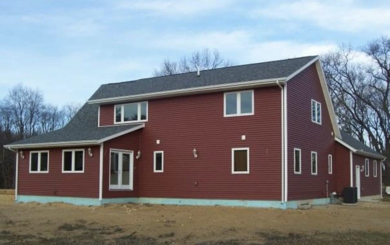 Red farmhouse-style new build with vaulted entry, American flag, and mature trees on a manicured lawn